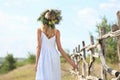 Young woman wearing wreath made of beautiful flowers near wooden fence on sunny day, back view Royalty Free Stock Photo