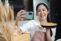 Young woman wears apron taking selfie photo with homemade pie in kitchen. Portrait of beautiful Asian female baking dessert and Royalty Free Stock Photo