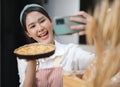 Young woman wears apron taking selfie photo with homemade pie in kitchen. Portrait of beautiful Asian female baking dessert and Royalty Free Stock Photo