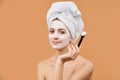 Young woman with white bath towel in her hair holding brush, applying face mask. Wellness and Spa concept on beige. Royalty Free Stock Photo