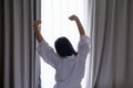 Young woman in a white bathrobe stretching in the morning against the background of window with open curtains.