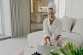 Young woman in white bathrobe working remotely on laptop while sitting on sofa in modern living room Royalty Free Stock Photo