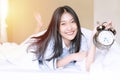 Young woman on white blanket holding alarm clock while lying on the white bed looking at camera. Cheerful girl waking up Royalty Free Stock Photo