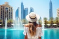 Young woman in white dress and hat on the background of Dubai fountain, Happy tourist girl rear view walking near fountains in