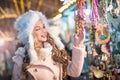 Young woman with white fur cap admiring accessories in Xmas market, cold winter evening. Beautiful blonde girl in winter clothes