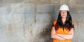 Young woman in white hard hat and orange high visibility vest, long dark hair, hands crossed, looking confident. Blurred Royalty Free Stock Photo