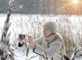 Young woman in a white jacket photographs winter canes of the forest lake coast on the phone