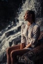 Young woman in white shirt and bikini sits on rock near waterfal Royalty Free Stock Photo