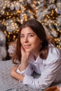 A young woman in a white shirt and blue jeans lies on a bed against a background of Christmas trees and bokeh of garlands Royalty Free Stock Photo