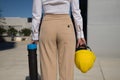 Young woman in white shirt, brown pants, with yellow workman`s helmet and blueprint pipe in hands, back turned, arriving at offic