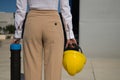 Young woman in white shirt, brown pants, with yellow workman`s helmet and blueprint pipe in hands, back turned, arriving at offic