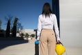 Young woman in white shirt, brown pants, with yellow workman`s helmet and blueprint pipe in hands, back turned, arriving at offic