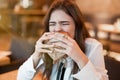 Young woman in white stylish blouse biting with appetite fresh meat burger during lunch in trendy resturant looking hungry eating
