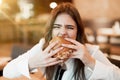 Young woman in white stylish blouse biting with appetite fresh tasty burger during lunch in trendy cafe feeling ultimate hunger