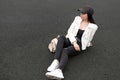 Young woman in white stylish leather jacket in a t-shirt in ripped jeans in white sneakers in a trendy baseball cap is resting Royalty Free Stock Photo