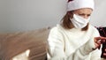 Young woman in a white sweater, red Santa Claus hat sits at home on the couch in a medical mask and prints a message on a mobile p