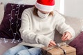 Young woman in a white sweater, Santa Claus hat and medical mask opens a New Year gift