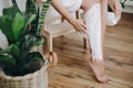 Young woman in white towel applying shaving cream on her legs in home bathroom with green plants. Skin care and wellness concept. Royalty Free Stock Photo