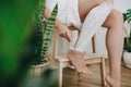 Young woman in white towel applying shaving cream on her legs in home bathroom with green plants. Skin care and wellness concept. Royalty Free Stock Photo