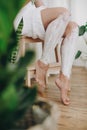 Young woman in white towel applying shaving cream on her legs in home bathroom with green plants. Skin care and wellness. Hair Royalty Free Stock Photo