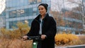 Young woman in winter black jacket with hat riding electic scooter in the city park. Ecological public transport