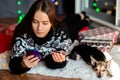 Young woman in winter sweater makes Christmas purchases with a small dog via phone at home online Royalty Free Stock Photo
