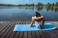 A young woman on a wooden dock sits on a blue mat in a fetal position with her arms around her legs. Meditation, yoga in nature Royalty Free Stock Photo