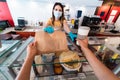 Young woman worker serving takeaway breakfast inside bar cafeteria while wearing face safety mask during coronavirus outbreak -