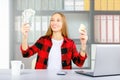 Young woman at workplace holding dollar bills and credit card, looking at money with smile.