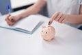 Young woman writing on notebook inserting coin on piggy bank at home Royalty Free Stock Photo