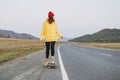 Young woman in yellow hoodie and red hat on skateboard on the road against beautiful mountain landscape, Chuysky tract, Altai
