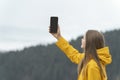 Young woman in yellow jacket in mountains holds phone in hand with blank empty black screen. Copy space. Mock up Royalty Free Stock Photo