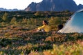 A young woman in yellow jacket sits next to a camping tent on the background of a beautiful landscape at sunset, traveling to