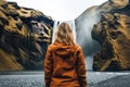 A young woman in a yellow jacket standing in front of a waterfall in Iceland, rear view of a Woman overlooking a waterfall at
