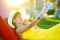 Young woman in yellow summer dress taking selfie with her mobile phone resting on a bench in park Royalty Free Stock Photo