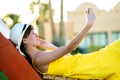 Young woman in yellow summer dress taking selfie with her mobile phone resting on a bench in park Royalty Free Stock Photo
