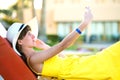 Young woman in yellow summer dress taking selfie with her mobile phone resting on a bench in park Royalty Free Stock Photo