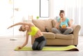 Young woman doing exercise on mat while her stout friend eating chips at home