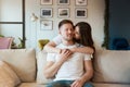 Young woman embracing her husband`s shoulders and kisses him while he sits on the sofa feeling love, romantic relationships