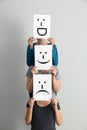 Young women hiding faces behind sheets of paper with drawn emoticons on light background