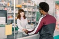 Young woman pharmacist giving medications to the male African client standing at the paydesk of the pharmacy store