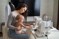 Young woman sews at home and holds a small child. Mom teaches her little son to sew on a sewing machine. Self-isolation Royalty Free Stock Photo