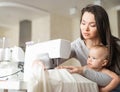 Young woman sews at home and holds a small child. Mom teaches her little son to sew on a sewing machine Royalty Free Stock Photo