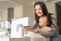 Young woman sews at home and holds a small child. Mom teaches her little son to sew on a sewing machine Royalty Free Stock Photo