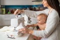 A young woman sews in the kitchen and holds a small child. Mom teaches her little son to sew on a sewing machine. Self Royalty Free Stock Photo