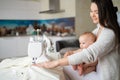 A young woman sews in the kitchen and holds a small child. Mom teaches her little son to sew on a sewing machine. Self Royalty Free Stock Photo