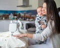 A young woman sews in the kitchen and holds a small child. Mom teaches her little son to sew on a sewing machine. Self Royalty Free Stock Photo