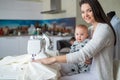 A young woman sews in the kitchen and holds a small child. Mom teaches her little son to sew on a sewing machine. Self Royalty Free Stock Photo