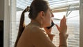 Young worried woman talking by phone and looking through window blinds. Crime witness, spying through window, peeking on street Royalty Free Stock Photo