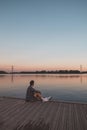 Young 25 year old brown man with plaid shirt sitting on the end of a wooden pier during sunset, watching the calm water surface in Royalty Free Stock Photo